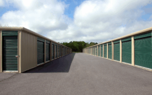 Exterior view of a modern self storage facility with long rows of green roll up doors on both sides of a paved drive aisle under a partly cloudy sky
